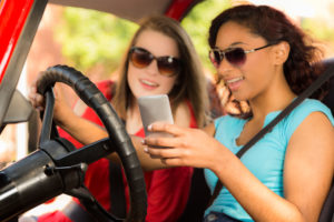 Texting and driving. Two teen girls text message on smart phone while driving their car. Mixed-race girl, caucasian girl friends.