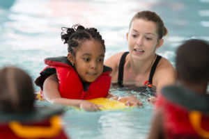 A multi-ethnic group of elementary age children are learning how to swim at the public pool. One little girl is holding onto a kick board and is swimming through the water.